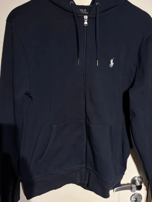 Mörkblå hoodie från Polo Ralph Lauren - Snygg mörkblå hoodie med dragkedja från Polo Ralph Lauren. Klassisk design med vit broderad logga på bröstet, huva med dragsko och två fickor framtill. Perfekt för en avslappnad och stilren look.