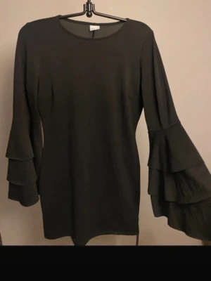 Svart klänning med volangärmar - Svart kortklänning med rund halsringning och långa ärmar med dubbla volanger. Klänningen har en enkel, rak passform och är tillverkad i ett mjukt material som ger en snygg siluett. Perfekt för dig som gillar stilrena plagg med en twist.