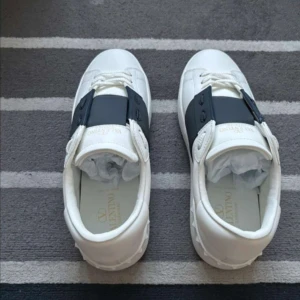 Vita Valentino sneakers med svart band - Säljer ett par vita Valentino sneakers med ett brett svart band över snörningen och ikoniska nitar på hälen. Skorna har rund tå och platt sula, perfekt för en stilren och modern look. Om de skulle va ngn fråga tveka inte på att skriva🫡