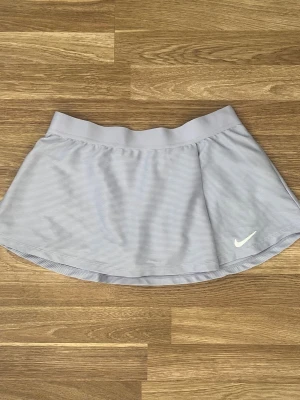 Ljuslila skort från Nike padel tennis kjol - Supersnygg ljuslila skort från Nike med diskreta ränder och klassisk vit logga framtill. Skorten har bred resår i midjan och är lätt utsvängd för en sportig look. Perfekt för dig som gillar en bekväm men trendig stil.
