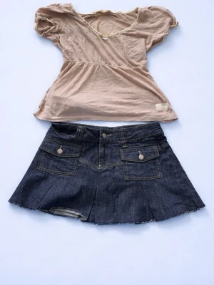 Mörkblå jeanskjol med fickor - Säljer en mörkblå jeanskjol i kort modell med två stora fickor framtill och dekorativa knappar. Kjolen har rå kant nertill för en avslappnad look och är sydd i kraftigt denimtyg. Perfekt för dig som gillar en trendig och casual stil.