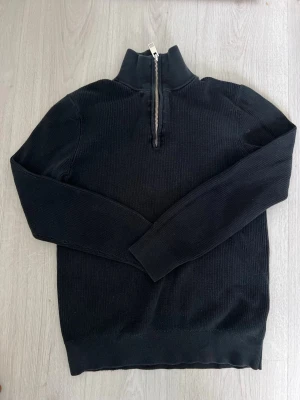 Svart quarterzip  - Snygg svart stickad tröja med hög krage och halv dragkedja framtill. Tröjan har lång ärm och ribbade muddar vid ärmslut och nederkant. Perfekt för lager på lager och enkel att matcha med olika outfits.
