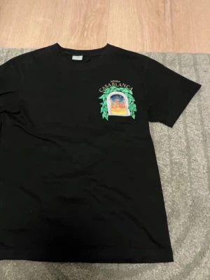 Svart Casablanca t-shirt med tryck - Säljer en svart t-shirt från Casablanca med färgglatt tryck av en byggnad och gröna blad både fram och bak. Texten 'Avenida Casablanca' syns tydligt. Klassisk passform och rund hals. Perfekt för dig som gillar streetwear och snygga prints.