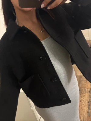 Svart croppad jacka från 157 - Stilren svart croppad jacka från 157 med klassisk krage och knäppning framtill. Jackan har raka fickor och är tillverkad i ett mjukt material som känns lyxigt. Perfekt för dig som gillar minimalistisk och trendig stil.