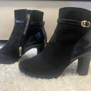 Svarta stövletter med krokodilmönster - Snygga svarta stövletter från Tamaris med klack och detaljer i krokodilmönstrat material. Dragkedja på insidan och guldfärgat spänne vid ankeln. Perfekta för att lyfta din outfit med en trendig touch.