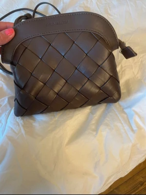  Ny Brun axelväska från Carin Wester - Snygg brun axelväska från Carin Wester i flätad läderdesign. Väskan har rundad form, justerbar axelrem och dragkedja upptill. Perfekt för dig som gillar stilrena detaljer och trendig look.