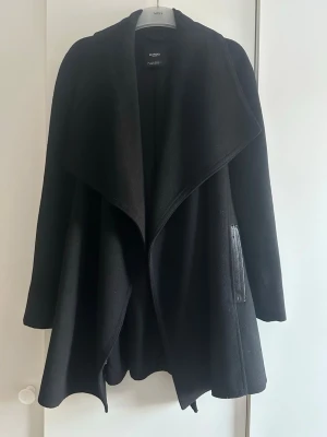 Svart kappa från Mango - Stilren svart kappa från Mango med bred krage och snygga detaljer i skinnimitation vid fickorna. Kappan har en avslappnad passform och är tillverkad i ett mjukt tyg som ger en lyxig känsla. Perfekt för dig som vill ha en trendig och tidlös look.