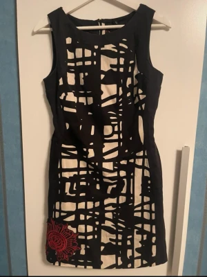 Svartvit kort klänning med röd brodyr - Unik kort klänning i svartvitt grafiskt mönster med en snygg röd broderad blomma vid nederkanten. Ärmlös modell med figurnära passform och rund halsringning. Perfekt för dig som vill sticka ut med en cool och modern look.