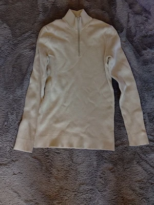 Beige ribbstickad tröja med halvzip - Snygg beige ribbstickad tröja med halvzip och hög krage. Tröjan har långa ärmar och är tillverkad i ett mjukt, stretchigt material som sitter skönt på kroppen. Perfekt för lager på lager och enkel att matcha med olika outfits.