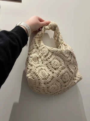Beige virkad handväska - Supersöt beige handväska i virkad design med rundad form och kort handtag. Väskan har ett unikt mönster med blommiga detaljer och stängs med dragkedja. Perfekt accessoar för en bohemisk look och rymmer det viktigaste du behöver.