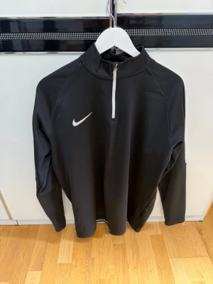 Svart Nike träningströja med dragkedja - Snygg svart långärmad träningströja från Nike med vit dragkedja och vit logga på bröstet. Tröjan har vita detaljer på axlarna och är tillverkad i ett stretchigt funktionsmaterial som passar perfekt till träning och sport.