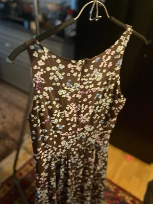 Blommig brun klänning  - Söt brun klänning med små blommor i blått, vitt och lila. Klänningen är ärmlös, har rund halsringning och dragkedja i ryggen. Perfekt för dig som gillar en feminin och lekfull stil.