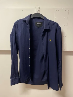 Mörkblå skjorta från Lyle & Scott - Stilren mörkblå skjorta från Lyle & Scott med klassisk krage och knappar framtill. Skjortan har lång ärm och den ikoniska gula loggan broderad på bröstet. Perfekt för en clean och enkel look. Slim Fit