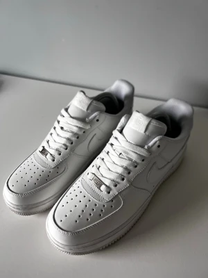Nike Air Force 1  - Klassiska Nike Air Force 1 sneakers i helvitt läder med perforerad tåbox, vita skosnören och den ikoniska swoosh-loggan på sidan. Tidlös design med låg profil och bekväm sula. Perfekta för dig som gillar clean och stilren look.