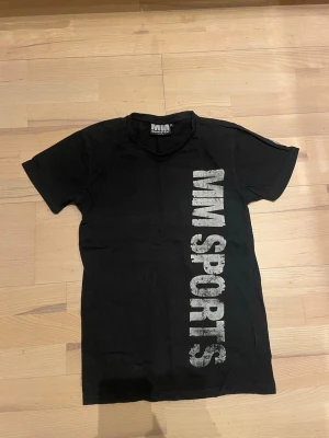 Svart MM Sports t-shirt - Säljer en svart t-shirt från MM Sports med stort tryck i vitt på både fram- och baksidan. Klassisk passform med rund hals och korta ärmar. Perfekt för dig som gillar sportig stil och vill ha något bekvämt till gymmet.
