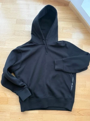 Svart hoodie från Calvin Klein Jeans - Svart oversize hoodie från Calvin Klein Jeans med klassisk huva och dragsko. Tillverkad i mjukt material med ribbade muddar vid ärmar och midja. Diskret logga tryckt på sidan. Perfekt för en chill och stilren look. Använd en gång pga fel storlek. 