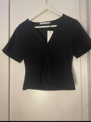 Svart croppad topp med knappar - Snygg svart croppad topp från Lager 157 med v-ringning och korta ärmar. Toppen har knappar framtill och en figurnära passform som ger en modern look. Perfekt att matcha med höga jeans eller kjol.