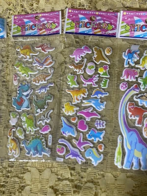 Färgglada dinosaurieklistermärken - Tre ark med klistermärken i olika färger och former, alla med dinosaurietema. Motiven inkluderar blå, gröna, gula och rosa dinosaurier samt några havsdjur och hjärtan. Perfekt för att dekorera anteckningsböcker, datorer eller pyssel.