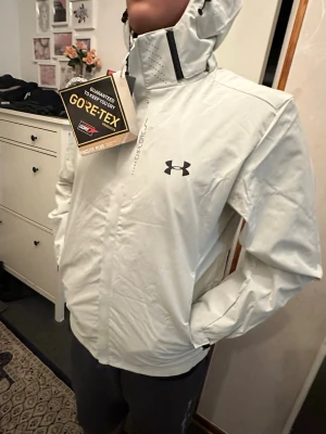 Vit vindjacka från Under Armour - Snygg vit vindjacka från Under Armour med GORE-TEX Paclite Plus-material som håller dig torr. Jackan har hög krage med ventilationshål, dragkedja framtill och två sidofickor. Under Armour-logga på bröstet och diskret 'Explore'-text vid dragkedjan.