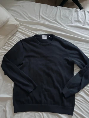 Mörkblå stickad tröja - En stilren blå stickad tröja med rund halsringning och långa ärmar. Tröjan har ribbade muddar vid ärmslut och nederkant, perfekt för en clean och enkel look. Passar till det mesta och är lätt att matcha med andra plagg.