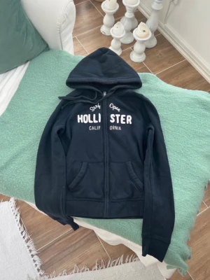 Svart hoodie från Hollister - Svart hoodie med dragkedja från Hollister. Stor vit brodyrtext på bröstet med 'Surf Open Hollister California'. Mjuk bomullsblandning, två fickor fram och justerbar huva med snören. Perfekt för en avslappnad och cool stil.