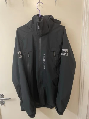 Svart vindjacka från Sail Racing - Svart vindjacka med huva från Sail Racing. Jackan har två vertikala bröstfickor med dragkedja, logga på bröstet och reflekterande tryck på båda ärmarna. Tillverkad i ett lätt och tåligt material som står emot vind. Perfekt för dig som vill ha en stilren och funktionell jacka.