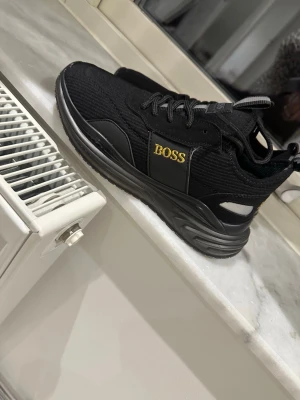 Svarta sneakers  - Snygga svarta sneakers  med gulddetalj på sidan och logga på plösen. Ovandelen är i textil med inslag av syntetmaterial och har svarta snören. Sulan är svart och har en chunky design som ger en modern look.