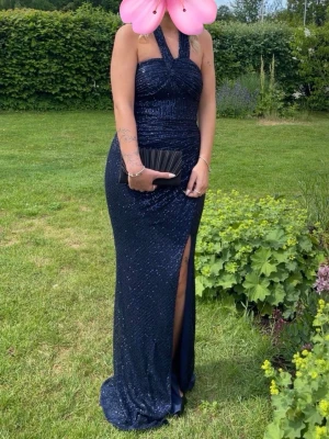 Balklänning Mörkblå glittrig  - Elegant mörkblå långklänning med glittriga paljetter, halterneck och hög slits framtill. Klänningen har figurnära passform och öppen rygg, perfekt för speciella tillfällen. Materialet är glittrigt och ger en lyxig känsla.
