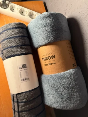 Två filtar i blå toner - Säljer två mysiga filtar, en enfärgad ljusblå i mjukt material (140x200 cm) och en blågrå med mörkblå ränder. Perfekta att ha i soffan eller sängen för extra komfort. Båda är lätta att rulla ihop och ta med.