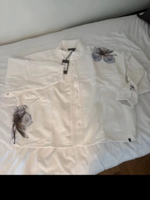 Vit skjorta med blommor från Elias rummelis - Vit skjorta från Elias rummelis med stora blommiga tryck i svart och blått på bröstet, ärmen och axeln. Skjortan har knappar framtill, klassisk krage och lång ärm. Tillverkad i bomull och har en loose passform för en avslappnad stil.