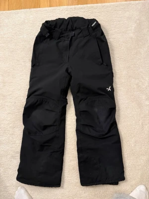Svarta täckbyxor från Everest - Säljer ett par svarta täckbyxor från Everest med justerbar midja, dragkedjefickor och förstärkta knän. Byxorna har reflexdetaljer baktill och kardborre vid benslut för bästa passform. Perfekta för kalla dagar i backen eller snölek.