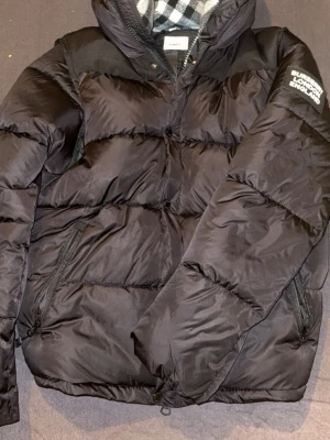 Svart pufferjacka från Burberry får en Lacoste armband med den  - Säljer en svart pufferjacka från Burberry med patch på ärmen där det står 'Burberry London England'. Jackan har dragkedja, knappar, två sidofickor och en huva med klassiskt rutigt foder. Perfekt för kalla vinterdagar och riktigt snygg streetstil. Kommer också med tag och brurbury påse 
