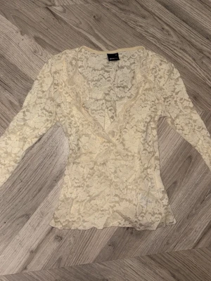 Krämvit spetsblus från Gina Tricot - Säljer en krämvit långärmad blus från Gina Tricot i genombruten spets med blommigt mönster. Omlott framtill med v-ringning och figurnära passform. Perfekt att styla med ett linne under för en trendig look.