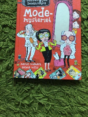 LasseMajas Detektivbyrå: Modemysteriet - En färgglad och rolig kapitelbok i serien LasseMajas Detektivbyrå, skriven av Martin Widmark och illustrerad av Helena Willis. Boken har ett rött omslag med tecknade figurer och handlar om ett mysterium i modevärlden. Perfekt för unga läsare som gillar spänning och humor.