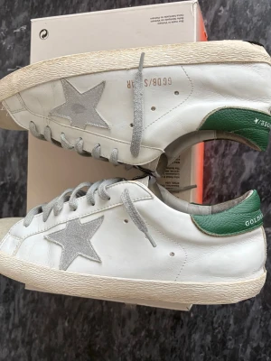 Golden Goose Superstar vita sneakers - Golden Goose Superstar sneakers i vitt läder med grå mockadetaljer och stjärna på sidan. Grön hälkappa med logga och grå skosnören. Klassisk låg modell med vintage-inspirerad sula och GGDB-märke på plösen. Perfekta för en trendig och avslappnad stil. Ny tvättade. Priset kan diskuteras