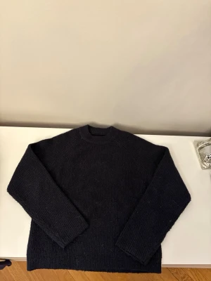 Svart stickad tröja med rund hals - En enkel och stilren svart stickad tröja med rund hals och långa ärmar. Tröjan har en rak passform och ribbstickade muddar vid ärmslut och nederkant. Perfekt för lager på lager och passar till det mesta.