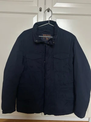 Mörkblå jacka från Woolrich - Snygg marinblå jacka från Woolrich med klassisk krage och fyra framfickor. Jackan har diskreta knappar och dragkedja framtill. Perfekt för dig som gillar stilrena och funktionella ytterplagg.