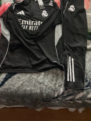 Real Madrid träningsset svart Adidas - Snyggt svart träningsset från Adidas med Real Madrid-logga och vita detaljer. Setet består av en långärmad tröja med halv dragkedja och matchande byxor med tre vita ränder på benet. Materialet är troligtvis polyester, perfekt för träning eller uppvärmning.