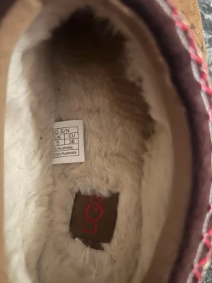 UGG tofflor med fårskinnsfoder - Mysiga tofflor från UGG i storlek 38 med mjukt vitt fårskinnsfoder och slitstark gummisula. Utsidan är beige med detaljer i rött och vitt. Perfekta för att hålla fötterna varma hemma under kalla dagar.