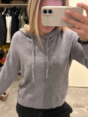 Grå hoodie med dragsko - Mysig grå hoodie med huva och dragsko i halsen. Tillverkad i mjukt stickat material som ger en avslappnad och stilren look. Ribbstickade muddar vid ärmslut och nederkant. Perfekt för en chill vardagsstil.