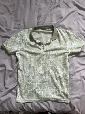 Grön mönstrad pikétröja från D designer  - Snygg ljusgrön pikétröja från Di designer med diskret logomönster över hela plagget. Tröjan har klassisk krage i en mörkare grön nyans och korta ärmar. Perfekt för dig som gillar exklusiva och trendiga plagg med unik design.