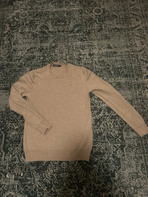 Beige stickad tröja J.Lindeberg - Snygg beige stickad tröja från J.Lindeberg med rund halsringning och långa ärmar. Tröjan är i ett tunt, mjukt material som passar perfekt till lager-på-lager. Enkel och stilren design som funkar till det mesta.