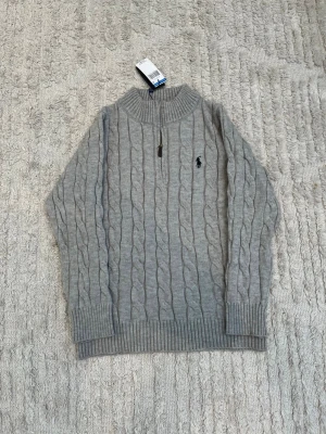 Grå kabelstickad tröja Polo Ralph Lauren - Kabelstickad grå tröja från Polo Ralph Lauren med halv dragkedja och hög krage. Tröjan har ribbade muddar och nederkant samt den klassiska broderade loggan på bröstet. Perfekt för lager-på-lager och snygg till jeans.