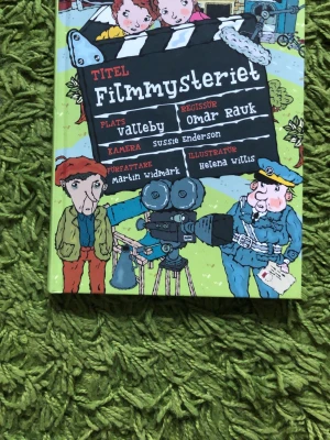 Filmmysteriet av Widmark & Willis - En färgglad och illustrerad bok ur LasseMajas Detektivbyrå-serie, skriven av Martin Widmark och illustrerad av Helena Willis. Omslaget har tecknade figurer, filmkamera och polismästare. Perfekt för dig som gillar spännande mysterier och roliga illustrationer.