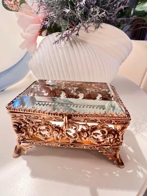 Vintage smyckeskrin i guld och glas - Unikt smyckeskrin i antik stil med guldfärgad metall och spegelglas på locket. Skrinet har vackra, detaljerade ornament och står på fyra dekorativa ben. Perfekt för att förvara smycken eller små accessoarer och blir en snygg inredningsdetalj.