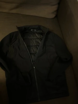 Svart jacka från Zara - Stilren svart jacka från Zara med dragkedja framtill och hög krage. Jackan har quiltad insida och är tillverkad i ett glansigt material som ger en modern känsla. Perfekt för dig som gillar enkel och clean design.