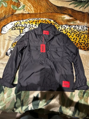 Svart overshirt från HUGO - Stilren svart overshirt från HUGO med två bröstfickor och röda logodetaljer på bröstet och insidan av kragen. Overshirten har klassisk krage, raka ärmar och är tillverkad i ett glansigt, lätt material som ger en modern look.