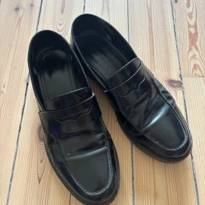 Svarta klassiska loafers i skinn - Snygga svarta loafers i glansigt skinn med klassisk penny loafer-design. Skorna har rund tå, platt sula och stilren look som passar till både jeans och kostymbyxor. Perfekta för dig som gillar tidlös och clean stil.