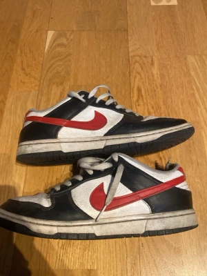 Nike Dunk Low svart/vit/röd - Nike Dunk Low sneakers i svart, vitt och rött med klassisk röd Swoosh på sidan. Ovandelen är i läder med perforerad tåbox och vita skosnören. Yttersulan är svart och mellansulan vit. Perfekt för dig som gillar retrostil och streetwear.