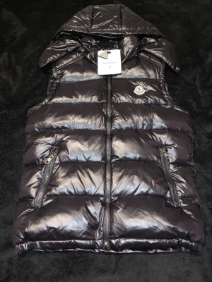 Svart dunväst från Moncler med huva - Snygg svart dunväst från Moncler med glansig finish och avtagbar huva. Västen har två dragkedjeförsedda fickor framtill och klassisk Moncler-logga på bröstet. Perfekt för lager-på-lager och streetstyle. Stängs med dragkedja framtill.